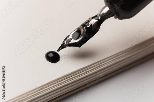 hand holds a smooth black pen in writing position against a clean white background where a drop of ink has just fallen. A contemporary ink drawing