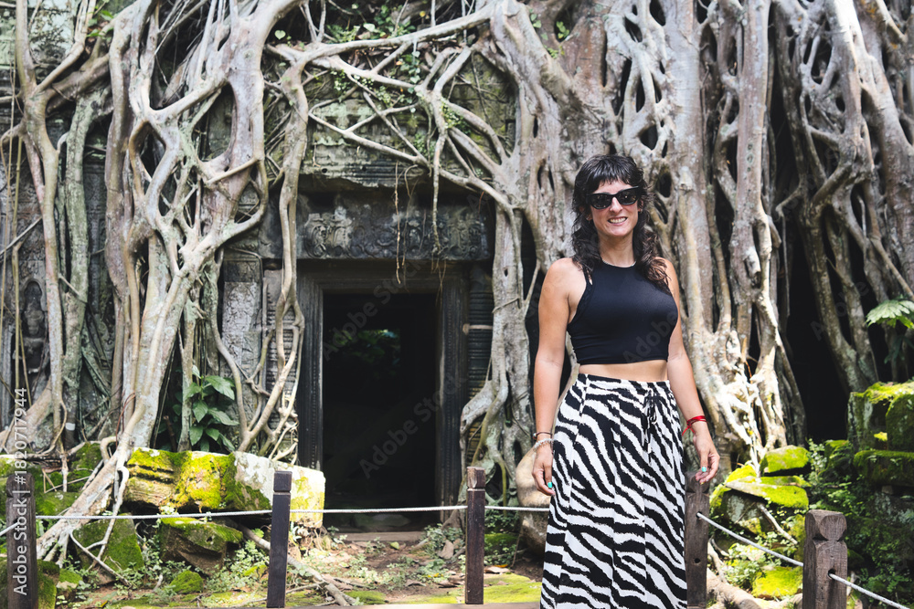 Obraz premium Young female traveler smiling beside ancient ta prohm temple, roots and jungle reclaiming mossy stone ruins in siem reap, cambodia exploration, history and adventure combined