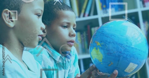 Examining two boys in light and blue plaid shirts, turning globe in classroom with shelf overlays