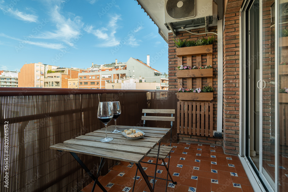 Obraz premium A cozy balcony in Barcelona with a wooden table and two glasses of wine. The view includes city buildings and a clear blue sky.