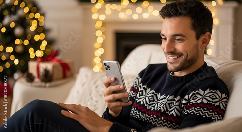 Happy man in a festive sweater looking at a mobile phone in a cozy living room decorated for Christmas holiday. Evening at home. © Iuliia