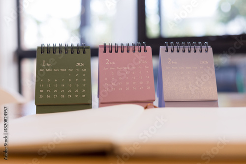 Colorful 2026 desk calendars showing January and February months on wooden table with blurred notebook ideal for planning and time management visuals.