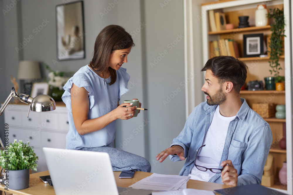 Obraz premium A woman sits on a desk, holding a coffee cup, engaging in conversation with a man who is seated in front of a laptop. They are in a cozy home office, effectively collaborating on their tasks.