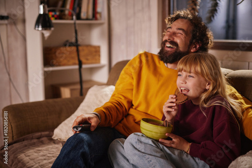 A man and a girl are sitting on a couch in a warm living room, laughing and watching a movie. They share snacks, creating a joyful atmosphere during their time together in the evening.