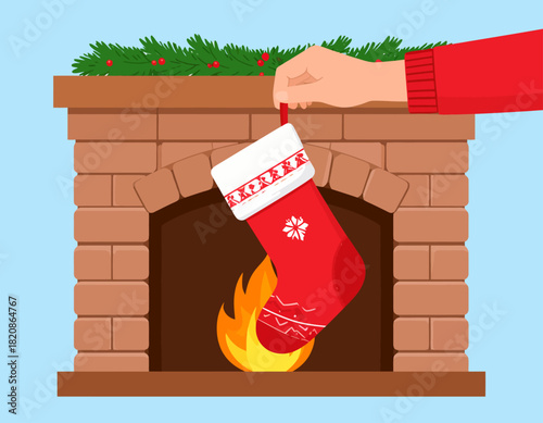 Hand hanging a red Christmas stocking with white snowflake patterns over a brick fireplace with a burning fire and green garland.