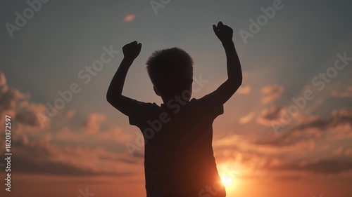 Child silhouette raises arms in sunset light. Boy celebrates freedom high arms in open sky. Arms lifted for victory. Sunset behind boy. Freedom moment for child with arms raised in sunset celebration.