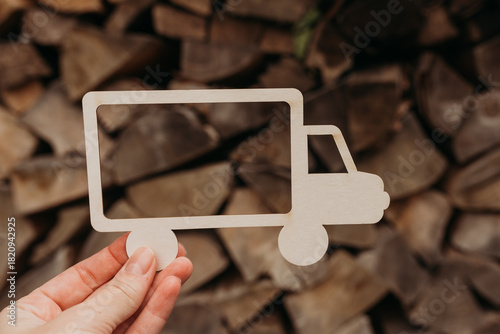 Hand holding a truck silhouette against a stack of firewood — wood delivery, forestry and logistics concept