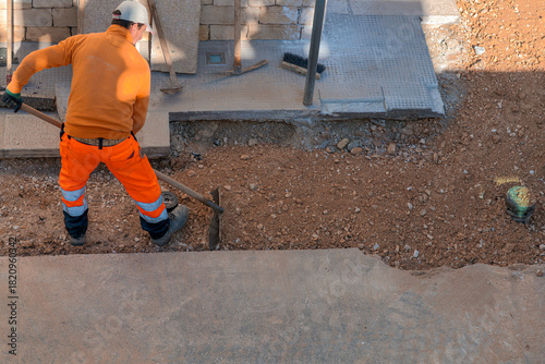 Top view of a construction worker in orange safety gear leveling gravel with a hoe. Manual labor and ground preparation at a building site during infrastructure renovation work.