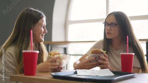 Girls share lunch at table eating burger and drinking soda from red cup tray in casual restaurant setting friend duo smile and talk each girl holds burger and enjoys meal together near bright window