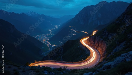 Winding mountain highway with car light trails at night. Long exposure captures traffic motion on serpentine pass. Illuminated village glows in valley below. Scenic roadtrip journey offers beautiful