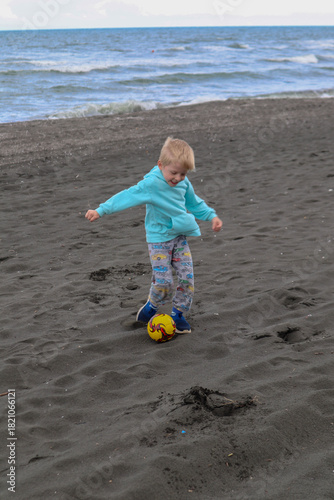 Little blond kid play football and have fun on black sand beach in Kobuleti, Georgia