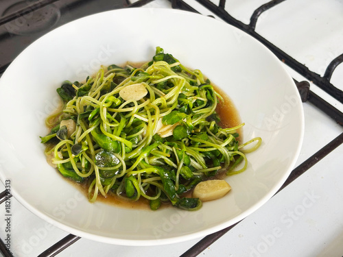 Close-up of sunflower sprouts stir-fried with oyster sauce, garnished with sliced garlic. The vibrant green color contrasts beautifully against the white plate, healthy cooking.