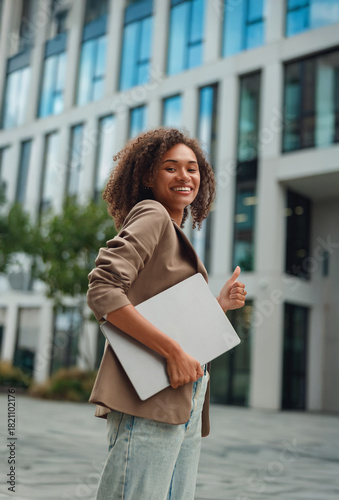 young black woman holding laptop confidently, walking toward office entrance, bright smile, professional