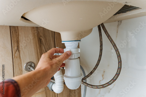 hand reaching under wooden sink basin to tighten trap and check connections, visible braided hoses and cabinet base, handson plumbing maintenance scene for home repair and service