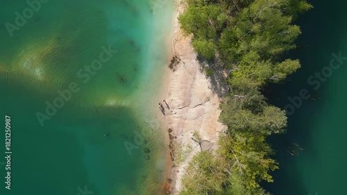 A bright sandy and rocky peninsula extends into turquoise water, surrounded by dense forest and clear underwater structures beneath the calm surface.