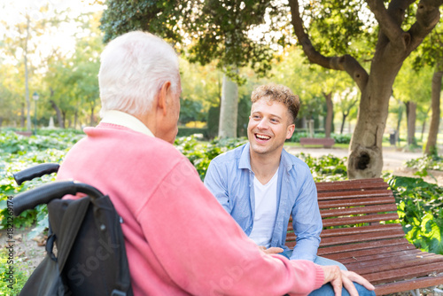 Young adult grandson sitting on bench talking and smiling with grandfather in wheelchair in park