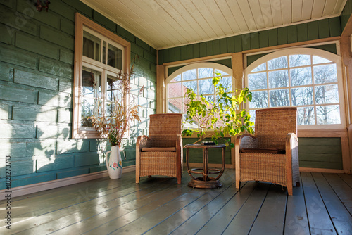 Billede på lærred Two wicker armchairs and a round table sit on plank flooring in an enclosed porch with teal paneled walls and arched multi pane windows casting crisp shadows in late autumn