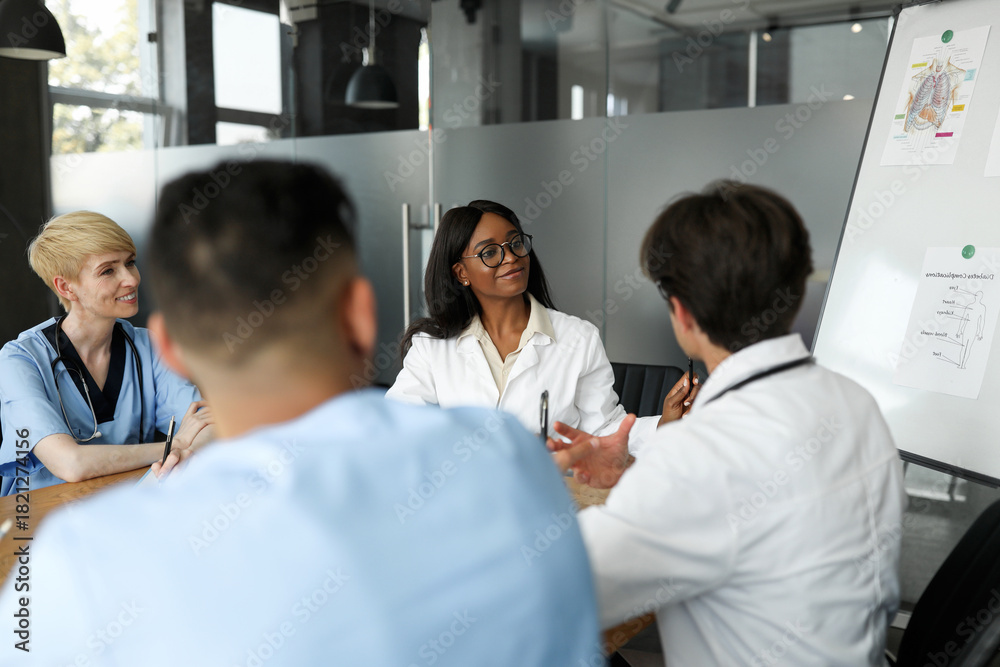 Fototapeta premium Multiethnic team of doctors having team-building, male and female physicians sitting at clinic, attending training, looking at presentation board and smiling, discussing new modern ways of treatment