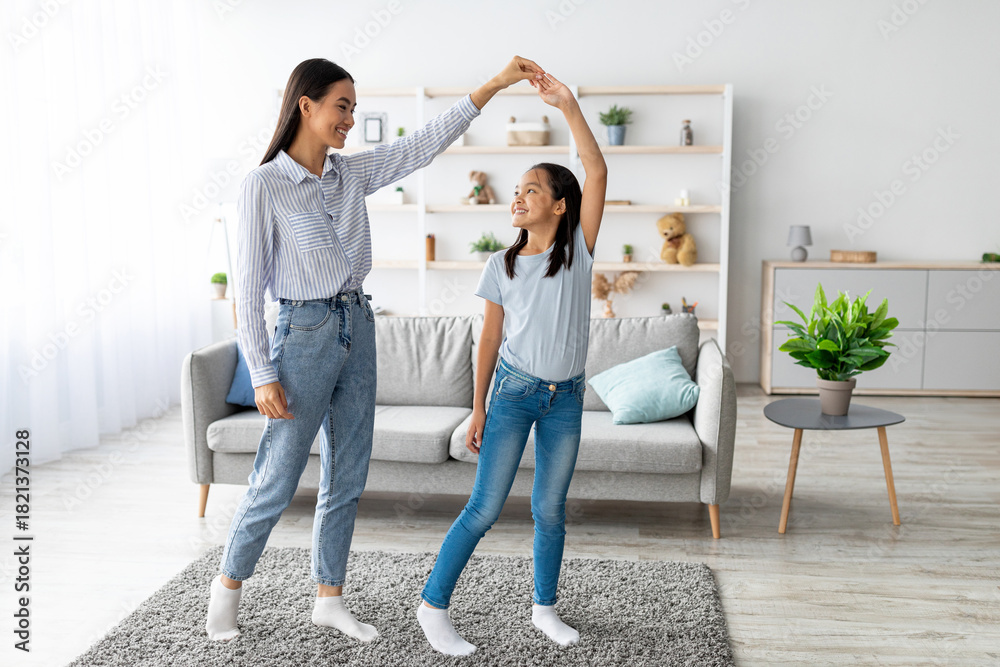 Fototapeta premium A cheerful mother and her daughter are dancing in their living room, holding hands and smiling. They are enjoying their time together, embracing the joy of music and family moments.