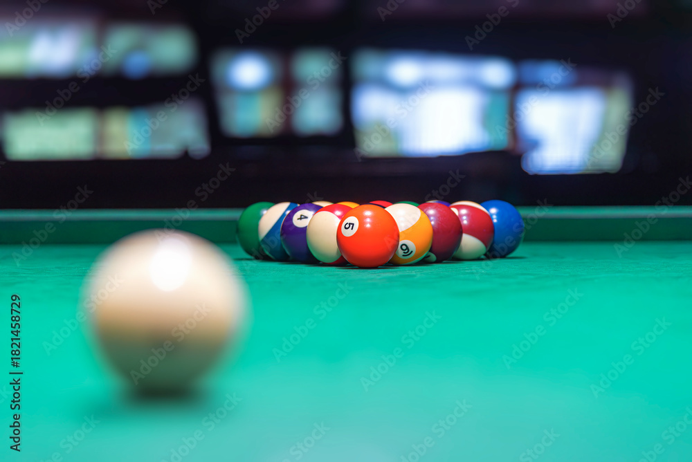 Fototapeta premium A single, white cue ball rests on a vibrant green pool table felt in a dimly lit setting, with a rack of colored billiard balls blurred in the background.