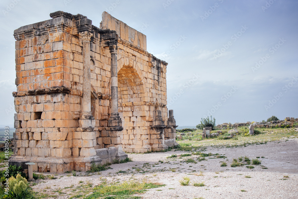 Fototapeta premium Ancient stone archway ruins stand majestically against a cloudy sky, surrounded by grassy terrain, showcasing historical architecture and remnants of a bygone era