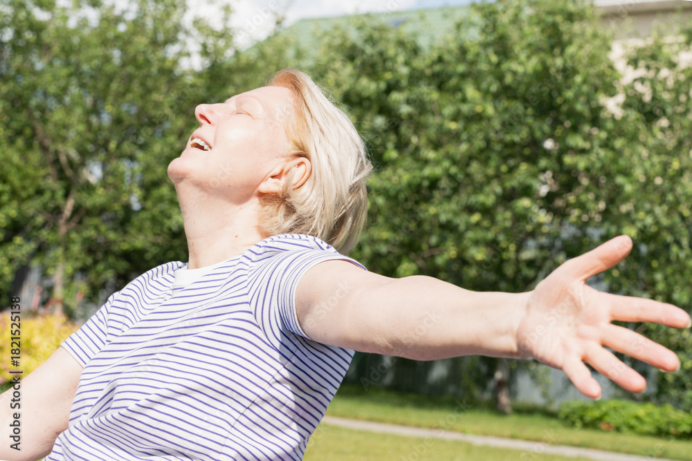 Obraz premium Senior woman smiling with eyes closed and arms wide open, enjoying sunshine and freedom in a garden