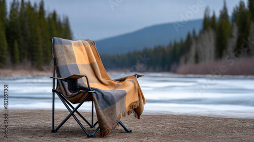 Winter riverside camp in forest with cozy chair and warm blanket near frozen water and pine trees