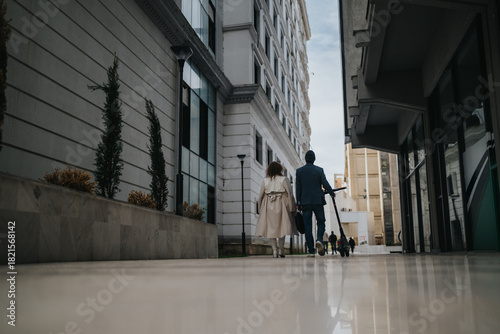 Wallpaper Mural A couple walks along a sleek city sidewalk, dressed in smart outfits, pulling a stroller as their child accompanies them. The urban backdrop features tall buildings and contemporary architecture. Torontodigital.ca