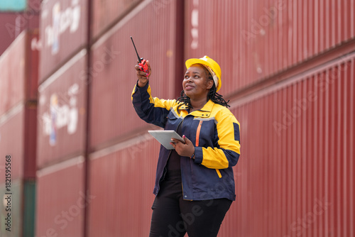 Confident female worker in safety gear uses walkie-talkie and tablet while overseeing shipping containers in logistics yard