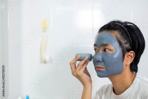 Asian woman applies clay facial mask in front of bathroom mirror. Skincare routine promotes clean skin and natural beauty. A refreshing beauty treatment for detoxifying and rejuvenating the face.