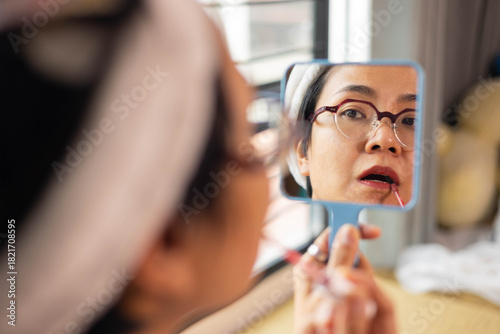Beautiful Asian woman applies lipstick in front of handheld mirror. Close-up reflection shows candid beauty routine of self care, natural skin, and confident expression during personal makeup moment.