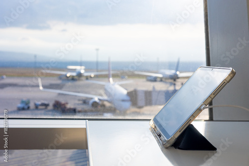 Selective focus on a tablet and smartphone charging by a sunlit airport window. In the blurred background, an airplane on the runway signifies travel, remote work, and the modern connectivity.