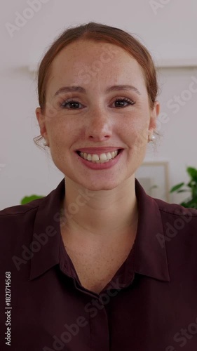 Vertical HD video. Portrait of young attractive businesswoman sitting on table while smiling at camera in the office. Close-up face portrait of beautiful ginger female manager. Webcam video call view.