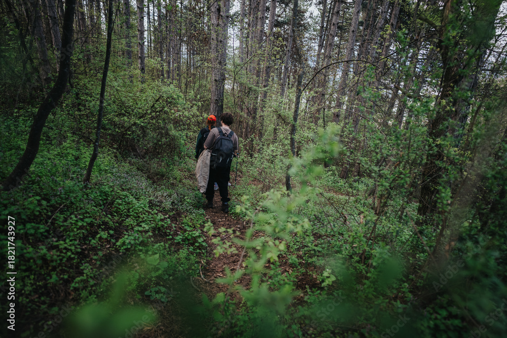 Fototapeta premium Two hikers backpacking through a dense green forest scene. The trail winds among trees and shrubbery, capturing a moment of exploration, nature, and calm adventure in a secluded woodland setting.