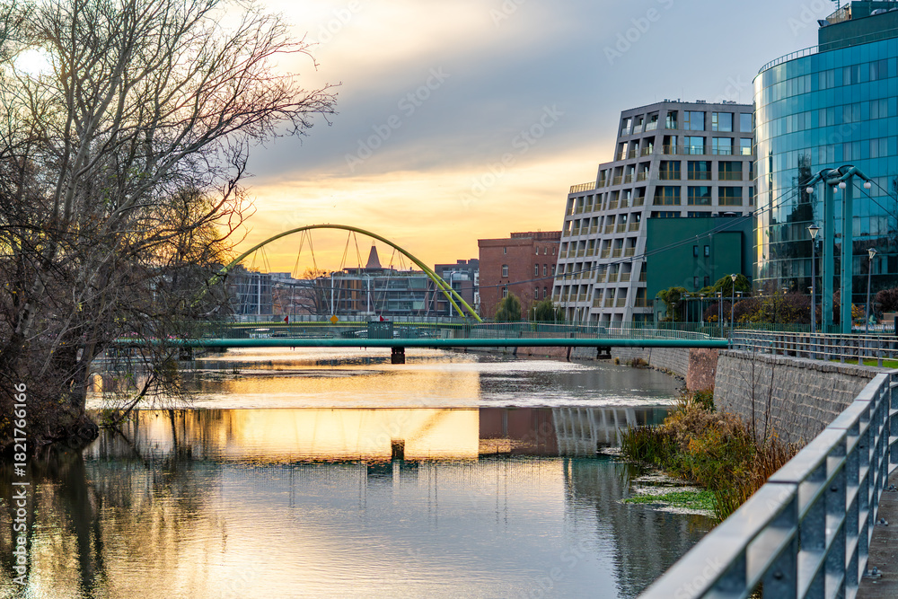 Obraz premium Wrocław skyline featuring Słodowa Footbridge over the Oder and modern office buildings reflecting in calm sunset water