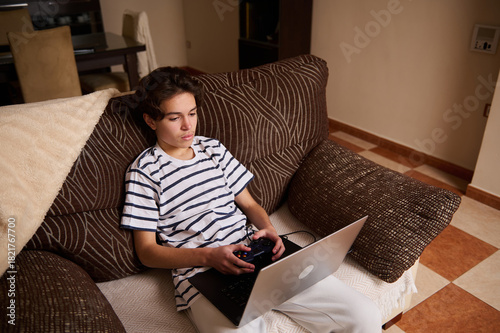 Teenage Boy Relaxing On Couch Playing Laptop Game Controller In Living Room