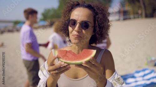 A young woman enjoying a slice of fresh watermelon on the beach while her friends relax and have fun in the background.