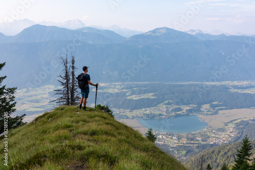 Billede på lærred A solo hiker with a backpack and poles stands on a grassy summit on Graslitzen mountain in Austria, contemplating the breathtaking panoramic view of Lake Pressegger and distant Carnic and Julian Alps