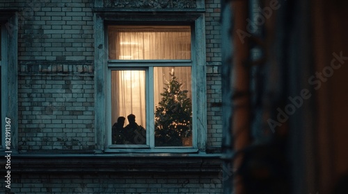 Fototapeta Naklejka Na Ścianę i Meble -  A view of a city apartment window from the street, showing the silhouette of people decorating their small tree inside,
