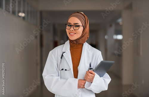 Portrait of happy Muslim female doctor or nurse wearing white coat with stethoscope in hospital corridor holding digital tablet looking at camera and smiling. Professional medical staff headshot.