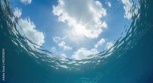 Fototapeta Naklejka Na Ścianę i Meble -  Underwater view looking up towards a bright sun and white fluffy cloud in a clear blue tropical ocean sky