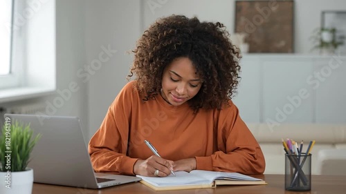 Happy Young African American Woman Writing in Notebook at Home Desk with Laptop, Engaged in Study or Remote Work, Creative Process, Learning, or Ta...