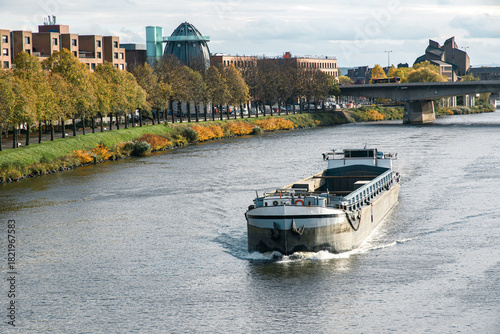 Maastricht Netherlands cargo barge sails on the River Maas past autumn trees modern buildings and road bridge urban waterway logistics inland shipping sustainable transport European city travel scene