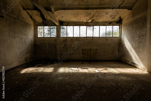 interno fatiscente di un edificio abbandonato. 
con pareti di cemento grezzo e un pavimento sporco e disordinato e conluce naturale che filtra attraverso piccole finestre