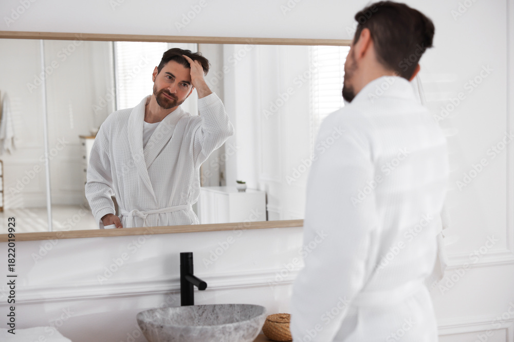Fototapeta premium Handsome man wearing bathrobe and looking at himself in mirror indoors
