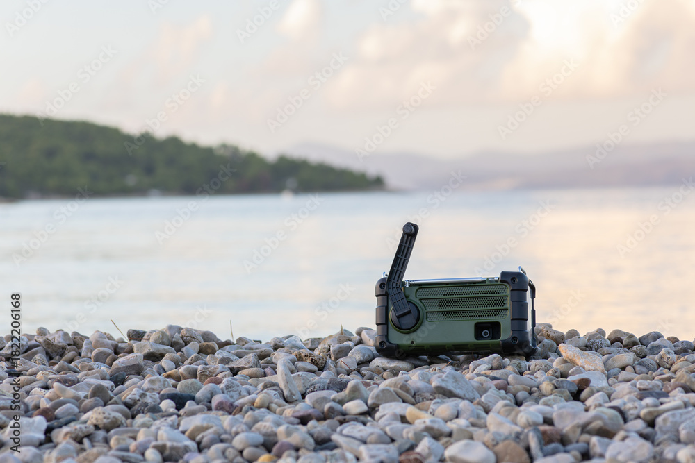 Fototapeta premium Rugged Green Radio Resting On Pebble Shore By Calm Sea, Outdoor Communication Gear