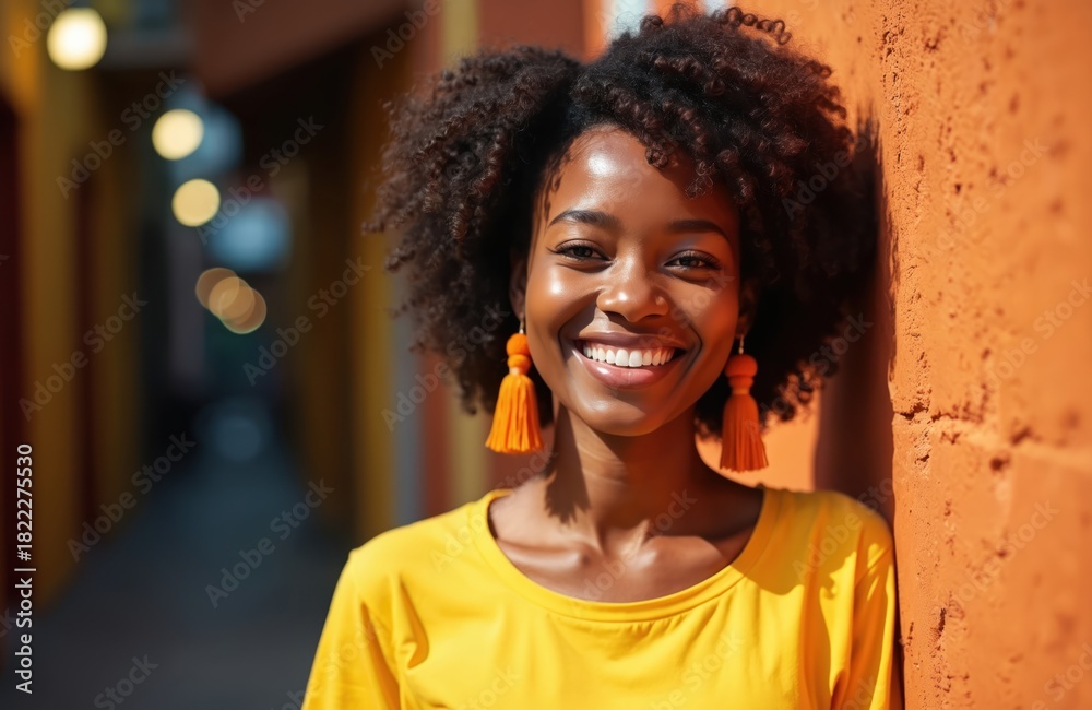 Fototapeta premium Black woman smiles near orange wall. She wears yellow shirt and big earrings. Curly afro hair. Joyful person on a sunny day. Portrait photo.