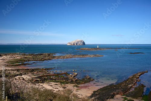 A tranquil coastal scene at Seacliff Beach with Bass Rock in the distance. Blue water, rocky shoreline and a distant lighthouse capture East Lothian's rugged Scottish coast.
