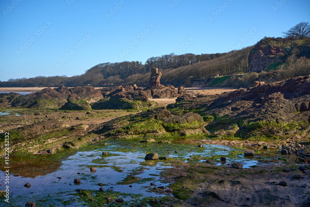 Obraz premium Waves recede on a rugged shore at Seacliff Beach in East Lothian, Scotland, revealing tidal pools and mossy rocks beneath a bright sky.