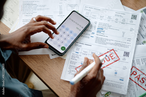 Black young man analyzing invoices and bills using smartphone calculator and pen, hands holding pen over documents with overdue and past due stamps on wooden desk